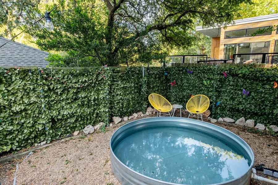 Backyard stock-tank pool at 1413 Luna Street after grading — clear blue-green water, saturated ivy screen, yellow acacia chairs in late-afternoon light.