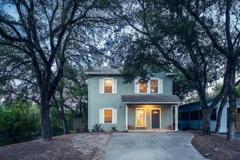 Two-storey pale-green house at blue hour, windows glowing warm under live oak branches.