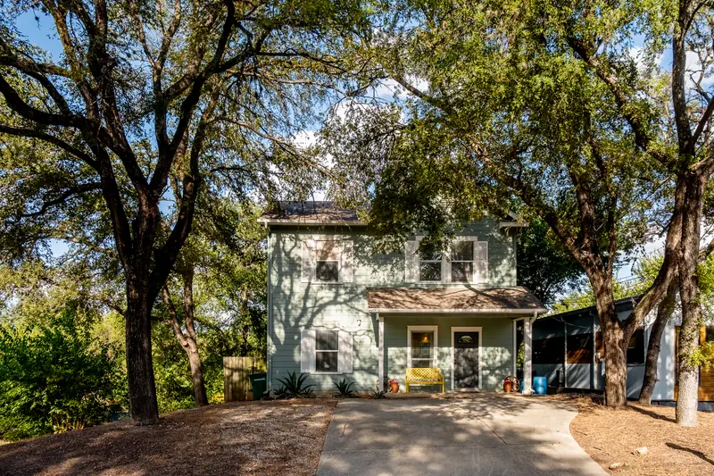 Front elevation of a pale-green two-storey house framed by live oaks in afternoon light.