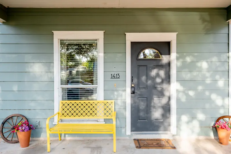 Covered front porch with a yellow metal bench, potted flowers and a grey front door.