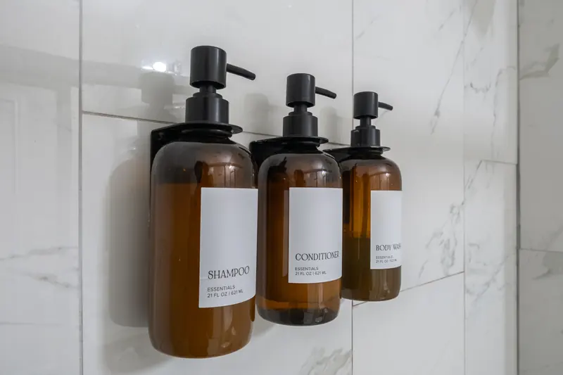 Three refillable amber amenity bottles mounted on a marble shower wall.