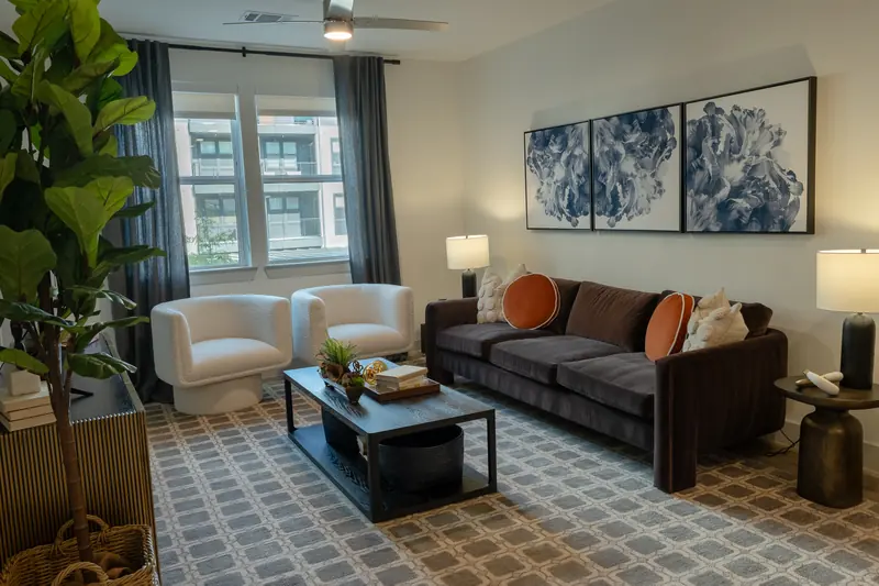 Model apartment living room — white armchairs, dark sofa, blue diptych artwork and a fiddle-leaf fig.