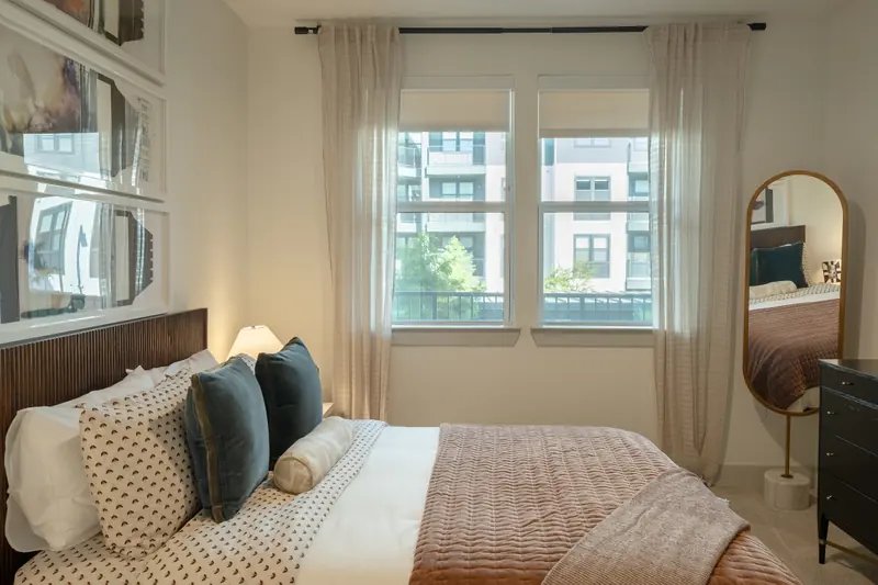 Model apartment bedroom with a rust throw, framed prints above the bed and window light.