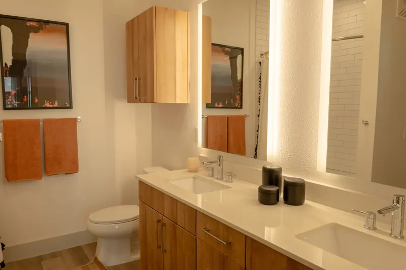 Double vanity with a backlit mirror niche, wood cabinetry and orange towels.